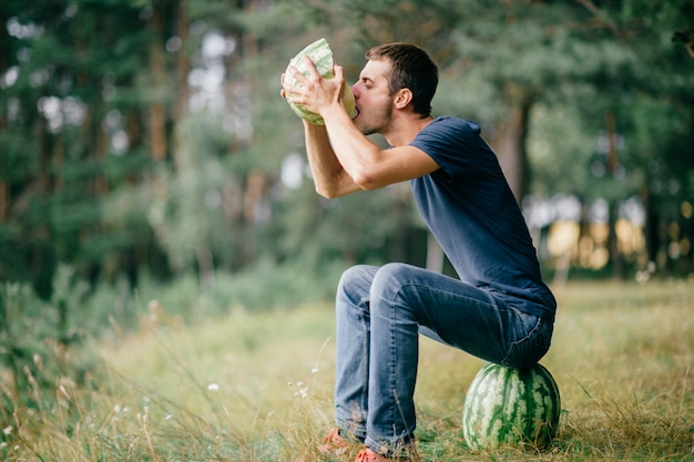 Jeune homme étrange assis sur une pastèque, la main tenant ses mains et buvant son jus