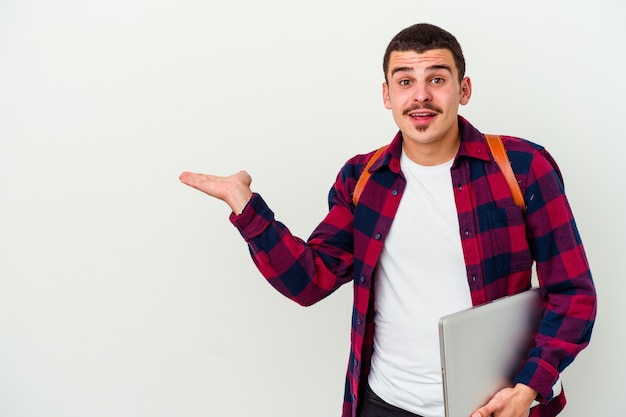 Jeune homme étudiant caucasien tenant un ordinateur portable isolé sur un mur blanc détient un espace de copie sur une paume, garder la main sur la joue