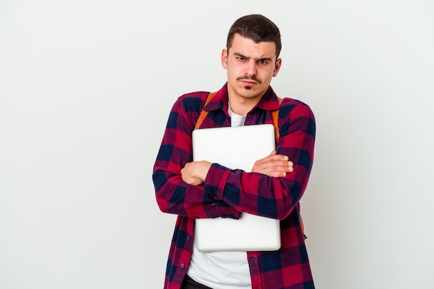 Jeune homme étudiant caucasien tenant un ordinateur portable isolé sur un mur blanc fronçant le visage de mécontentement, garde les bras croisés.