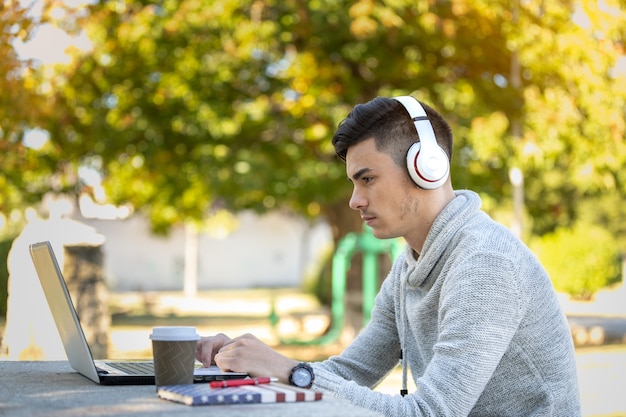 Jeune homme étudiant avec ordinateur tout en écoutant de la musique avec des écouteurs dans le parc sans masque