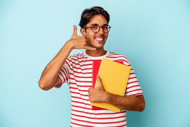 Jeune homme étudiant de race mixte tenant des livres isolés sur fond bleu montrant un geste d'appel de téléphone portable avec les doigts.