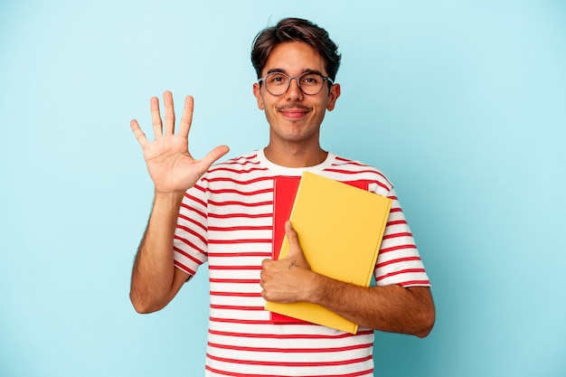 Jeune homme étudiant de race mixte tenant des livres isolés sur fond bleu souriant joyeux montrant le numéro cinq avec les doigts.