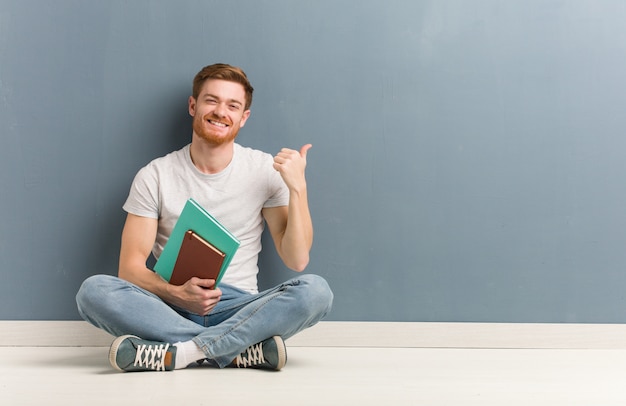 Jeune homme étudiant rousse assis sur le sol pointant vers le côté avec le doigt Il tient des livres.