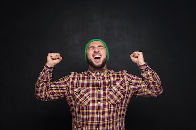 Photo jeune homme européen avec barbe en bonnet vert, a l'air surpris et perplexe. montrant les doigts vers le haut et le côté droit. fond noir, espace copie vierge pour le texte ou la publicité