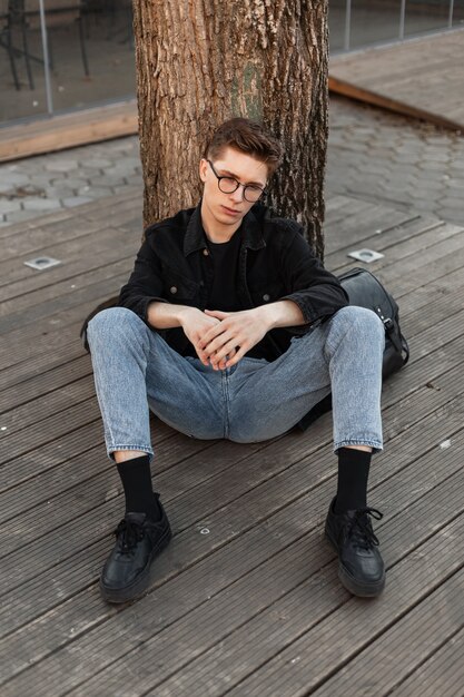 Jeune homme européen à la mode dans des lunettes vintage dans des vêtements en denim décontractés à la mode dans des baskets élégantes