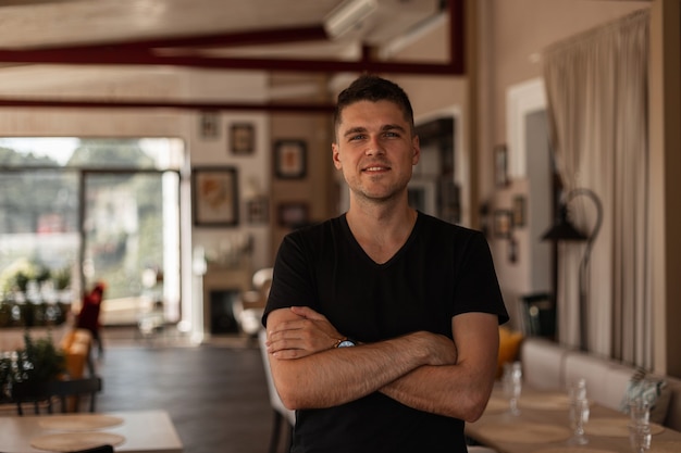 Jeune homme européen urbain avec une coiffure à la mode dans un élégant t-shirt noir se dresse et sourit dans un café vintage