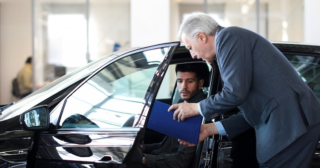 Jeune homme examinant une nouvelle voiture dans une salle d&#39;exposition pendant que le concessionnaire explique ses caractéristiques