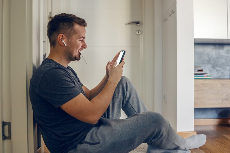 Un jeune homme excité est assis sur le sol et utilise un téléphone avec un écran tactile et tient la carte dans sa bouche