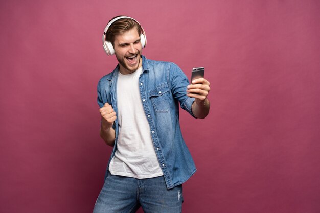 Jeune homme excité portant une chemise en jean isolé sur fond rose, écoutant de la musique avec des écouteurs et un téléphone portable