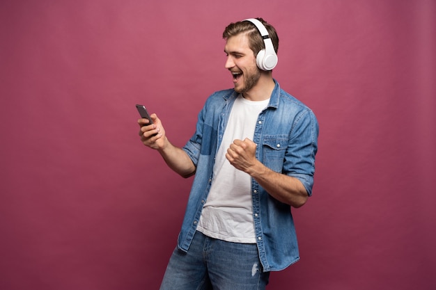 Jeune homme excité portant une chemise en jean isolé sur fond rose, écoutant de la musique avec des écouteurs et un téléphone portable