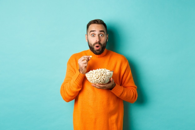 Jeune homme excité regardant un film intéressant sur un écran de télévision, mangeant du pop-corn et ayant l'air étonné, fond bleu