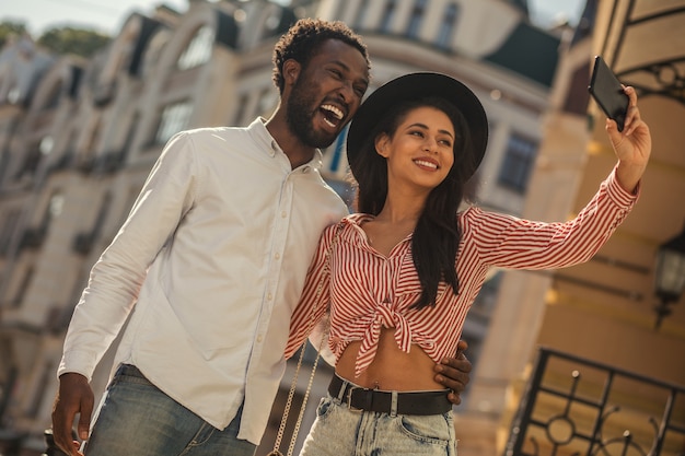 Jeune homme excité riant la bouche ouverte tandis que la jeune femme au chapeau prend un selfie avec lui