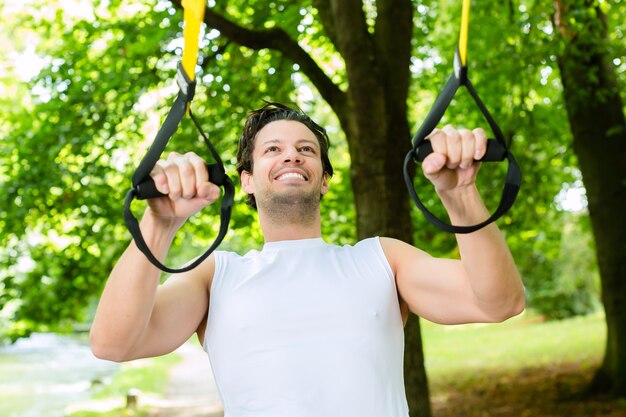 Jeune homme exerçant avec élingue de formateur de suspension dans le parc de la ville sous les arbres d'été pour le sport