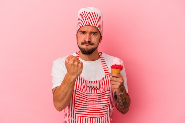 Jeune homme de fabrication de glace caucasien avec des tatouages tenant une crème glacée isolée sur fond rose montrant le poing à la caméra, expression faciale agressive.