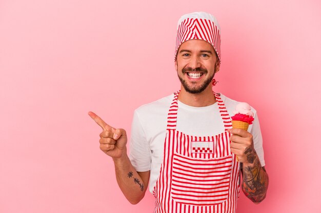 Jeune homme de fabrication de glace caucasien avec des tatouages tenant une crème glacée isolée sur fond rose souriant et pointant de côté, montrant quelque chose dans un espace vide.