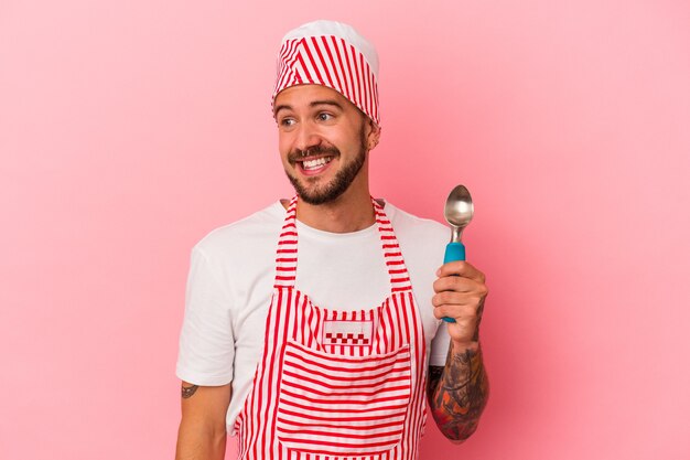 Jeune homme de fabrication de glace caucasien avec des tatouages tenant une cuillère isolée sur fond rose regarde de côté souriant, gai et agréable.