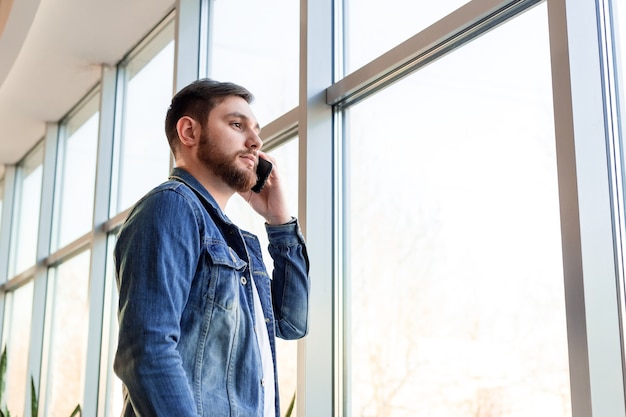 Jeune homme faisant un appel d'affaires parlant par téléphone à l'intérieur près du mur de la fenêtre dans une veste en jean décontractée. L'homme intelligent dans le bureau de la ville moderne a une conversation mobile. Homme d'affaires barbu du Caucase.