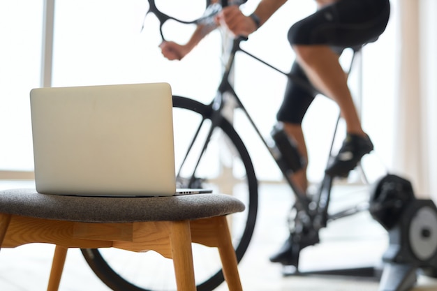 Jeune homme faisant du vélo stationnaire dans la chambre avec ordinateur portable