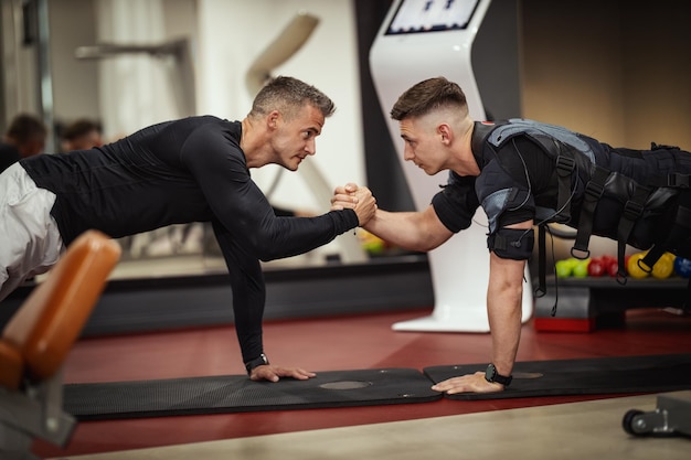 Un jeune homme fait des exercices de planche avec un entraîneur personnel dans la salle de gym, vêtu d'un costume noir avec un simulateur électronique EMS pour stimuler ses muscles.