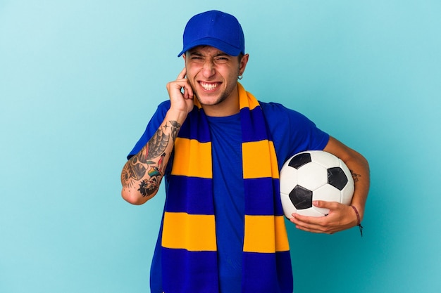 Jeune homme fan de football tenant un ballon isolé sur fond bleu couvrant les oreilles avec les mains.