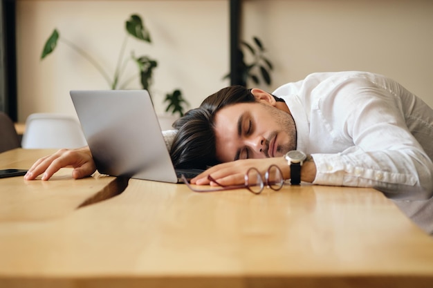 Jeune homme fatigué avec un ordinateur portable dormant sur le bureau au travail dans un bureau moderne