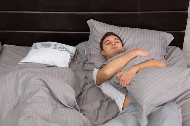 Jeune homme fatigué en pyjama dormant seul sans couverture et serrant un oreiller dans un lit élégant aux couleurs grises et près d'une table de chevet avec des bougies dans une chambre de style loft