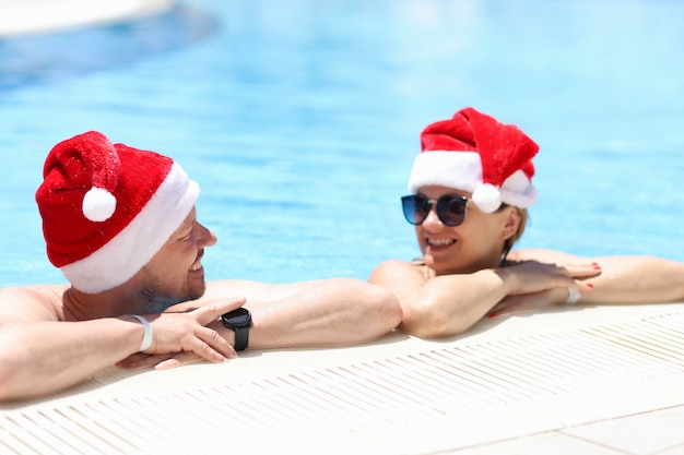 Jeune homme et femme à lunettes de soleil et chapeaux de père noël parlant dans la piscine