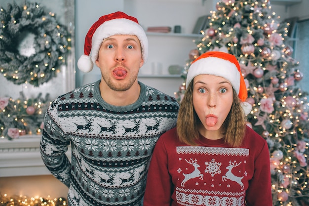 Jeune homme et femme en vêtements de fête regardent la caméra. Ils se tiennent ensemble et montrent leurs langues. Guy garde les mains derrière. Ils portent des chapeaux de Noël.