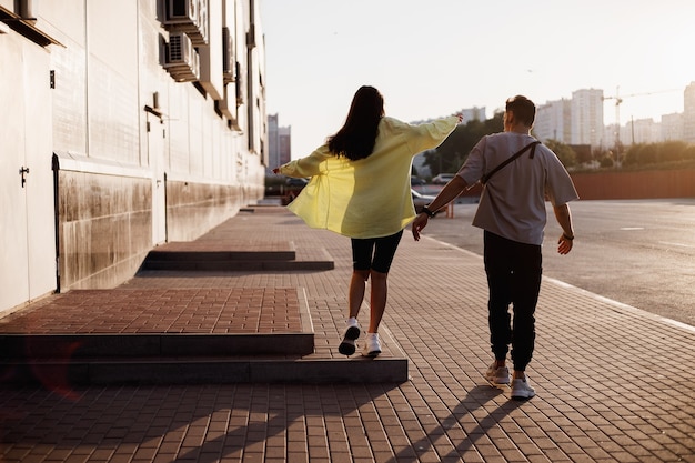 Un jeune homme et une fille élégants marchent ensemble sur la place pour se garer à côté du bâtiment au coucher du soleil.