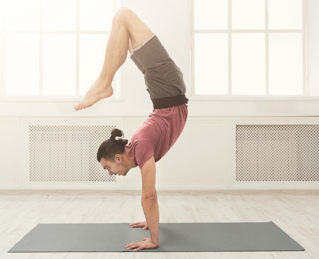 Jeune homme flexible pratiquant le yoga, debout sur les mains sur un tapis en cours de fitness, faisant des exercices d'équilibre, espace de copie