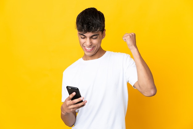 Jeune homme sur fond jaune isolé à l'aide de téléphone mobile et faisant le geste de la victoire
