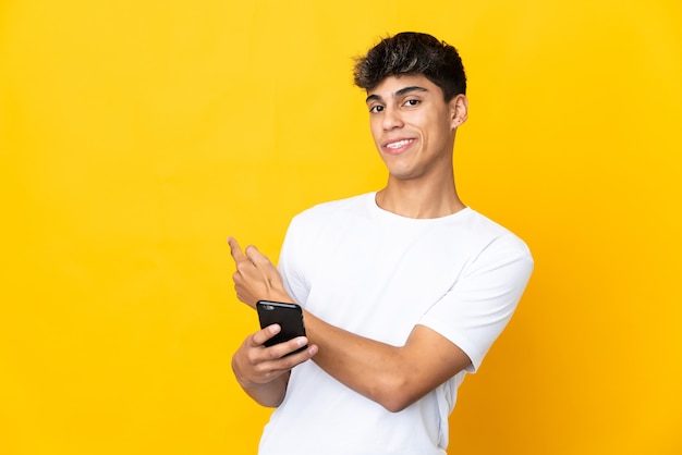 Jeune homme sur fond jaune isolé à l'aide de téléphone mobile et pointant vers l'arrière