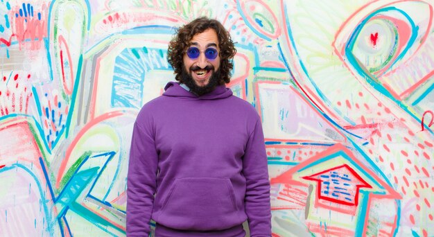 Jeune homme fou barbu à l'air heureux et maladroit avec un large sourire amusant et loufoque et des yeux grands ouverts contre le mur de graffitis