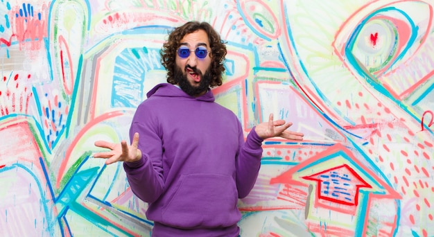 Jeune homme fou barbu, haussant les épaules avec une expression stupide, folle, confuse, perplexe, se sentant ennuyé et désemparé sur un mur de graffitis