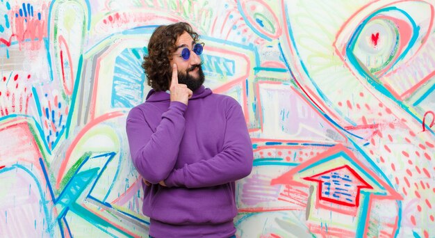 Jeune homme fou barbu avec un regard concentré, se demandant avec une expression douteuse, levant les yeux et à côté contre le mur de graffitis