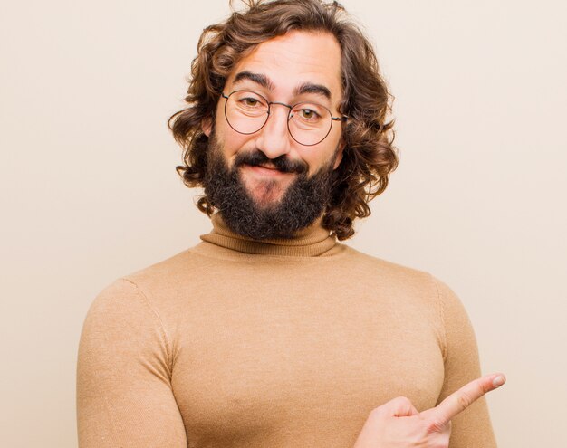 Jeune homme fou barbu souriant gaiement, se sentant heureux et pointant vers le haut et vers le haut, montrant un objet en fond contre une couleur plate