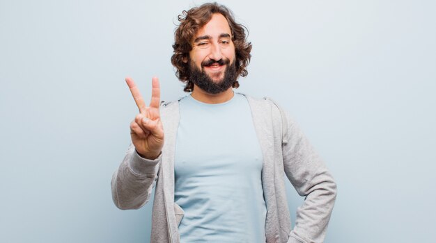 Jeune homme fou barbu souriant et à la recherche amicale, montrant le numéro deux ou seconde avec la main en avant, compte à rebours contre mur couleur plat