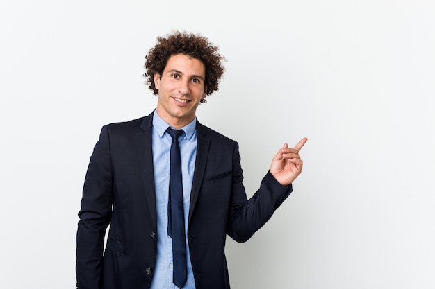 Jeune homme frisé d'affaires contre le mur blanc souriant joyeusement pointant avec l'index loin.