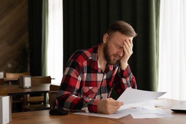 jeune homme frustré lisant des documents et utilisant un ordinateur portable dans un café problème d'argent notification de dette mauvais rapport financier problème d' argent résultats infructueux d' examens ou de tests