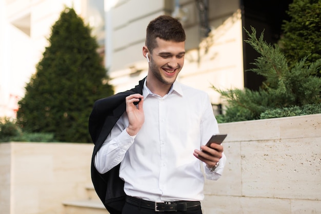 Jeune homme gai en chemise blanche avec des écouteurs sans fil tenant une veste noire sur l'épaule tout en utilisant joyeusement un téléphone portable en plein air