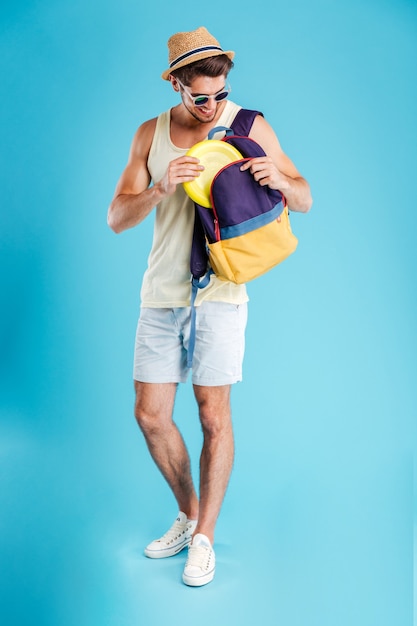 Jeune homme gai dans le chapeau et les lunettes de soleil prenant le disque de frisbee du sac à dos au-dessus du mur bleu