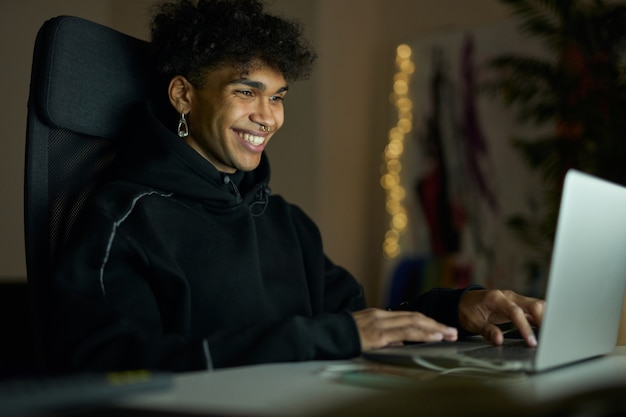 Jeune homme gai avec piercing souriant alors qu'il était assis devant l'écran d'un ordinateur portable à la fin du