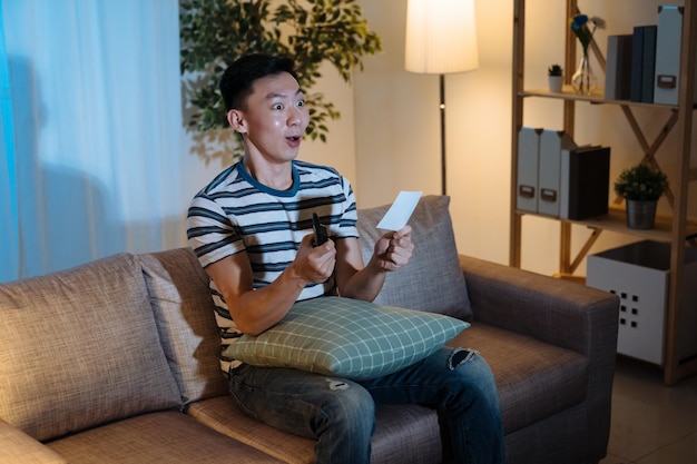 Jeune homme gai regardant un jeu de hasard à la télévision dans les mains de fin de nuit avec télécommande et billet de loterie assis sur un canapé et ayant l'air ravi. homme choqué surpris par la télé le soir