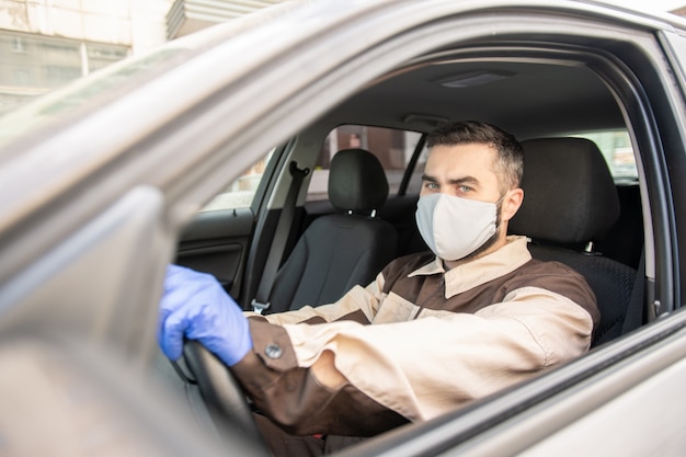 Jeune homme en gants de protection, masque et vêtements de travail assis dans la voiture et conduisant aux clients pour leur livrer des commandes en ligne