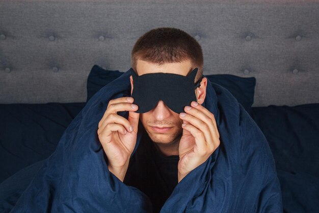 Un jeune homme gêné dans une couverture à la maison porte un masque de sommeil tout en se relaxant au home studio portrait Concept de style de vie relaxant dans la bonne humeur