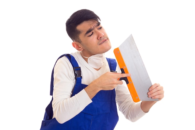 Jeune homme gentil aux cheveux noirs en chemise blanche et bleu dans l'ensemble avec respirateur blanc tenant une spatule sur fond blanc en studio