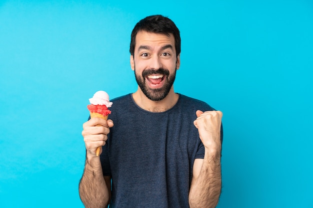 Jeune homme avec une glace au cornet sur un mur bleu isolé célébrant une victoire en position de gagnant