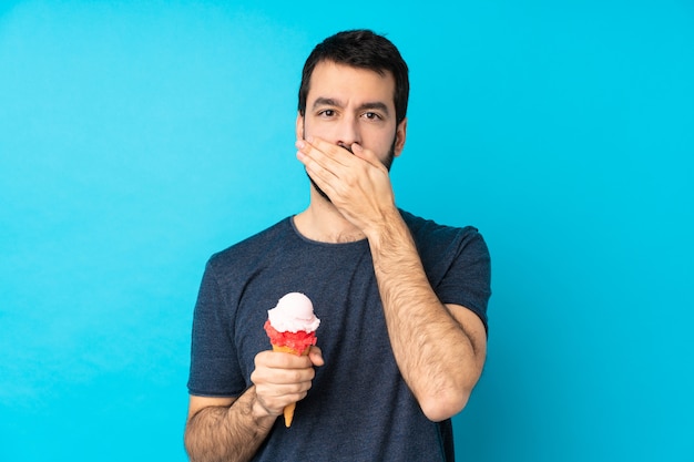 Jeune homme avec une glace au cornet sur un revêtement mural bleu isolé bouche avec les mains