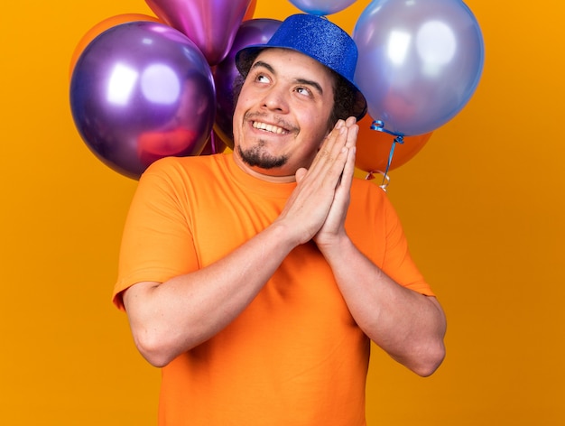 Jeune homme gourmand portant un chapeau de fête debout devant des ballons tenant la main ensemble isolé sur un mur orange