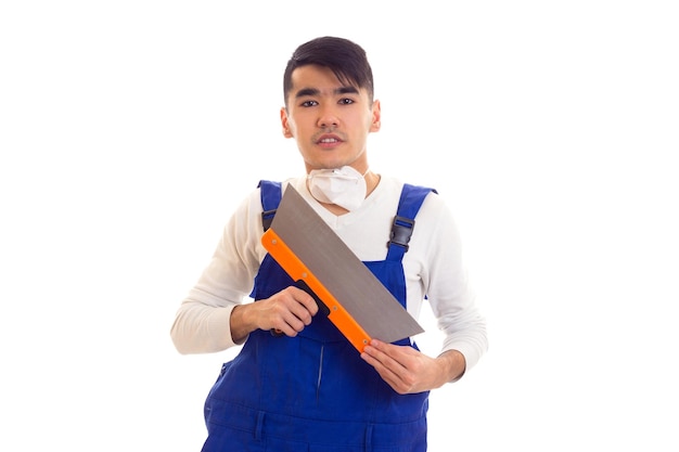 Jeune homme grave aux cheveux noirs en chemise blanche et combinaison bleue avec respirateur blanc tenant une spatule sur fond blanc en studio